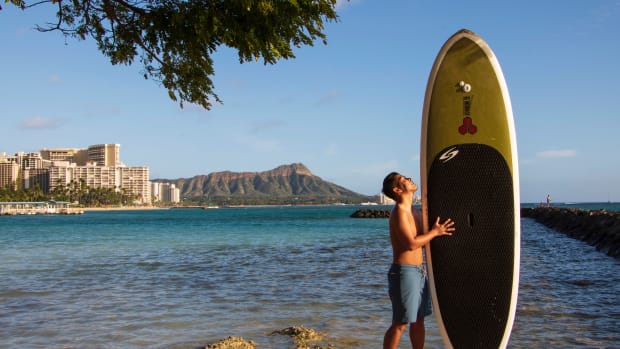 In this photo provided by Yoko Liriano, Bryant de Venecia poses for a photo with his paddleboard in Honolulu, Wednesday, Nov. 11, 2020. He started stand-up paddle-boarding when there were fewer tourists coming to Hawaii during the pandemic. He's among the Hawaii residents feeling ambivalence toward tourists returning now that the state is allowing incoming travelers to bypass a 14-day quarantine with a negative COVID-19 test. (Yoko Liriano via AP)