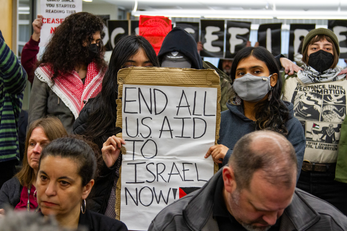 Two individuals hold up a piece of cardboard with signs on the front and back. One reads, “End all U.S. aid to Israel now.” (Photo by Jarrette Werk Underscore News/Report for America) 