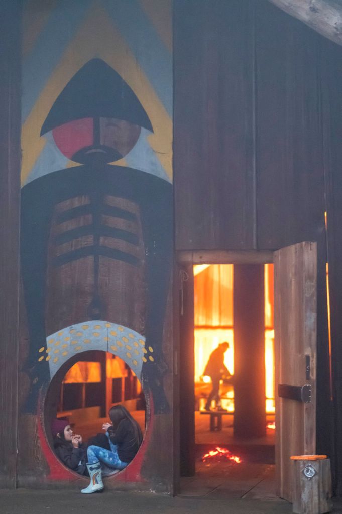 Two young people sit in a painted circular doorway of a Chinook plank house at the Ridgefield Wildlife Refuge, while warm firelight glows inside during the annual Winter Gathering.