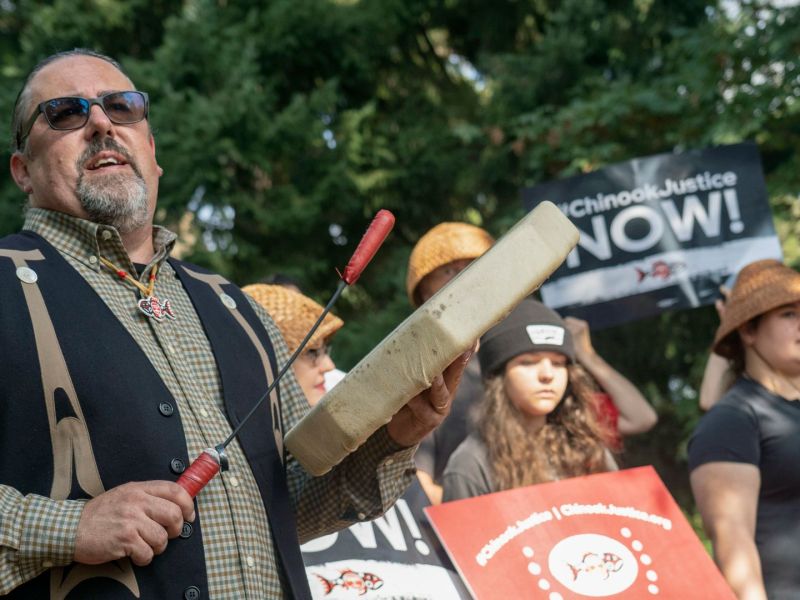 Chinook Indian Nation Chairman Tony Johnson sings and plays a hand drum during a 2025 rally for federal recognition, surrounded by supporters holding signs that read “#ChinookJustice NOW!” outside the Federal Offices in Vancouver, Washington.