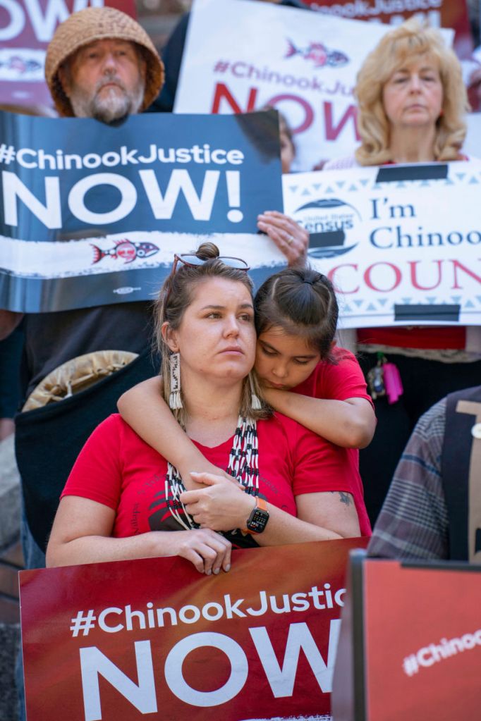 Chinook Indian Nation Council member Rachel Cushman holds a protest sign as her son Kanim, 7, hugs her from behind during a rally for federal recognition outside the Federal Courthouse in Seattle, surrounded by supporters holding signs reading “#ChinookJustice NOW!”