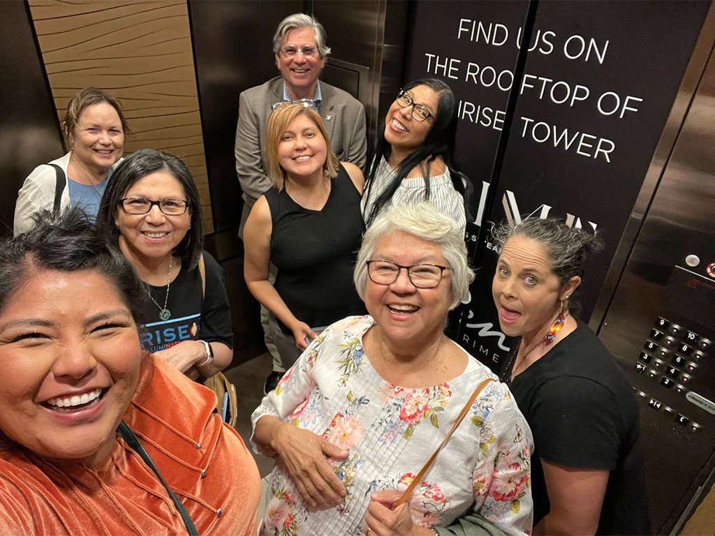 Jeanne Givens stands with ICT and IndiJ Public Media employees and board members inside an elevator in Phoenix, Arizona, smiling and posing for a group selfie in 2022.