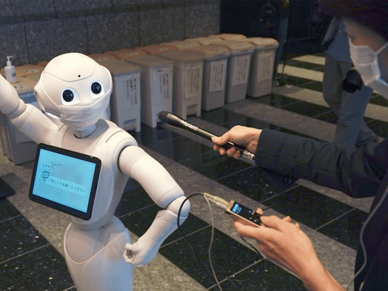 A humanoid robot wearing a face mask raises one arm while being interviewed by a masked reporter holding a microphone and audio recorder. The robot has a screen on its chest displaying Japanese text.