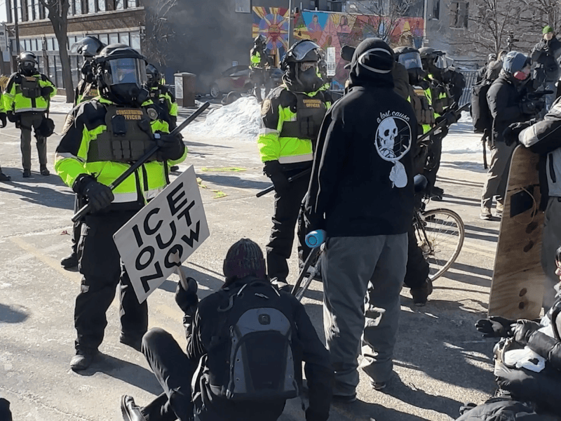 Protesters in Minneapolis, Minnesota stand and sit. Officers stand in opposition wearing gas masks, vests, and holding truncheons.