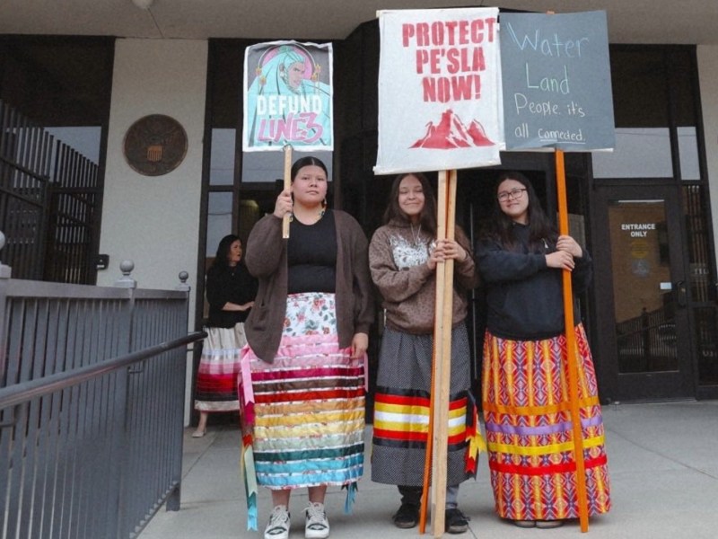 Indigenous women in ribbon skirts hold signs reading 'protect pesla now,' 'water, land, people: it's all connected, and 'Defund Line 3'
