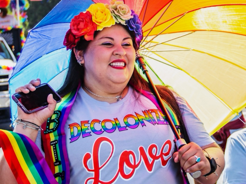 Smiling Indigenous woman celebrating PRIDE with rainbow colored flowers in hair and words Decolonize Love on shirt holding rainbow umbrella