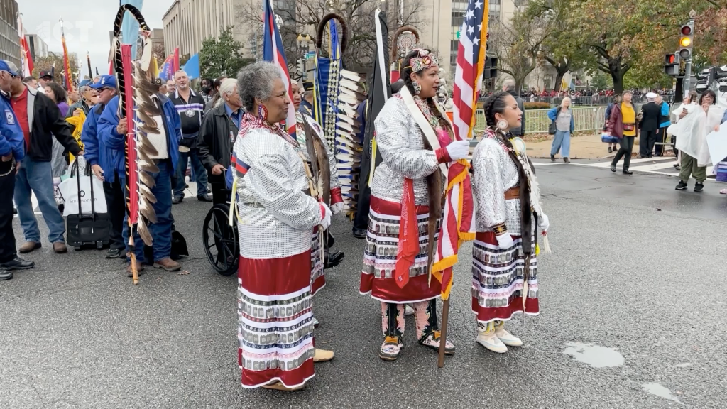Women in traditional Native American clothing stand near American flag