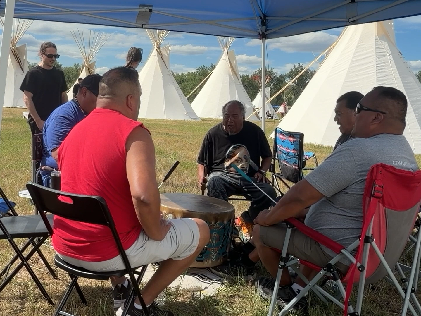 Native American men at Greasy Grass Celebrations sing and drum with teepees in background