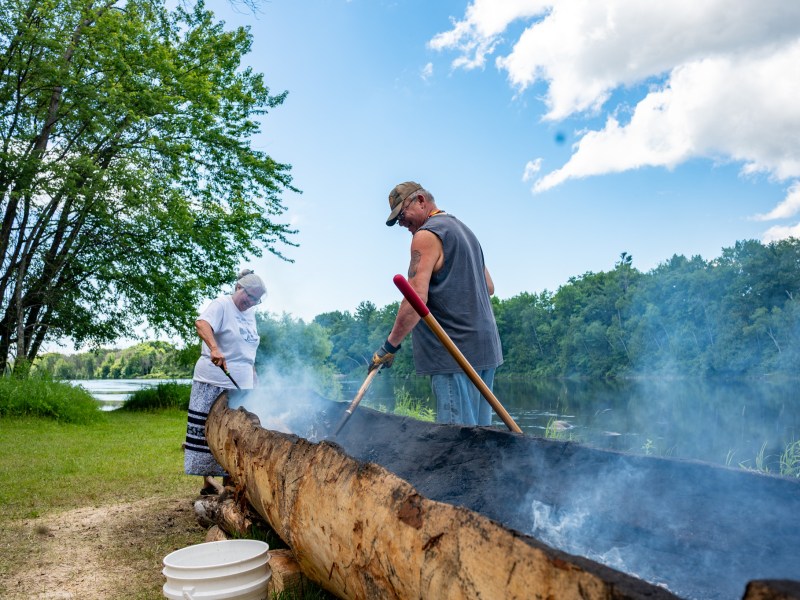 A man and woman stand around a burning pine log and work to chip away at the insides to create a canoe.
