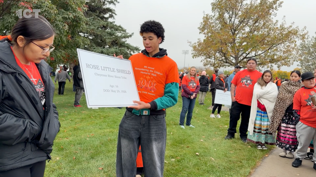 People gathered to remember the children who died at the Rapid City Indian Boarding School. A young man in an orange shirt holds sign with name "Rose Little Shield" Cheyenne River Sioux Tribe Age 16 DOD: May 29, 1926"